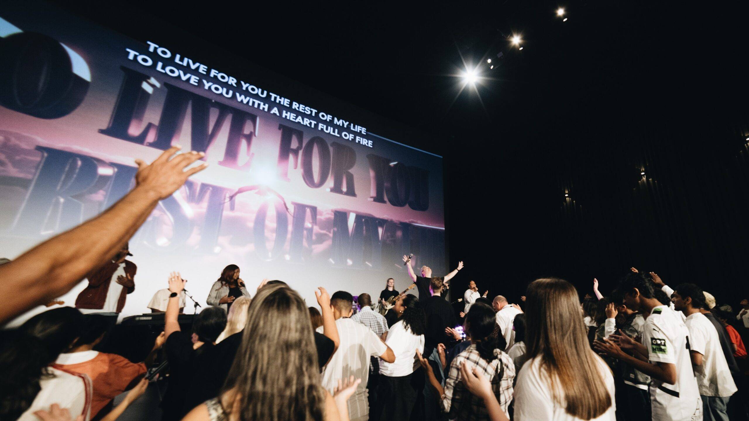 Crowd of people with raised hands at a worship concert in front of a large screen displaying a lyric text area behind the performers.