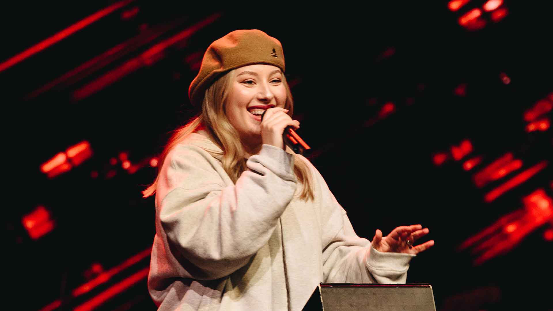 Young woman wearing a beige beanie and gray hoodie singing into a microphone on stage with red spotlights behind her.