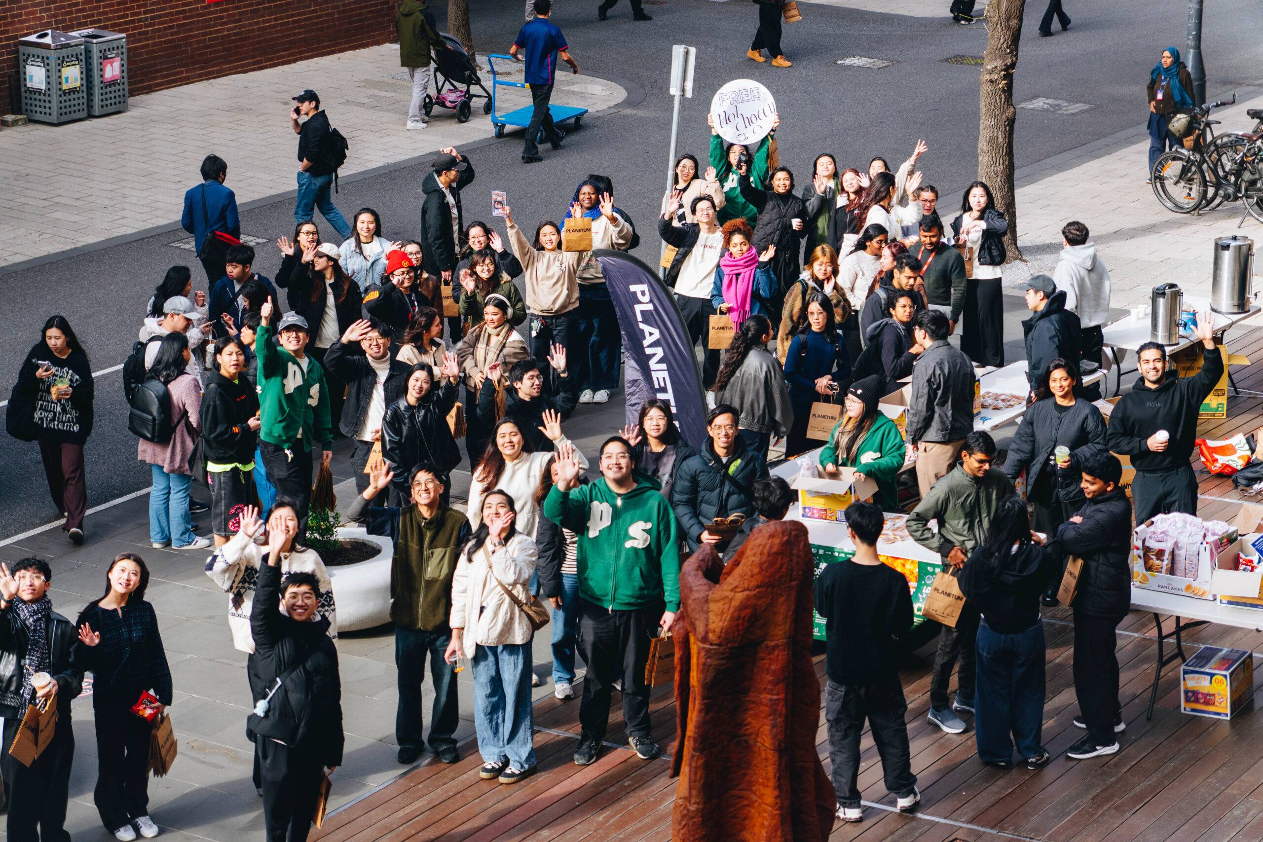 Large crowd of diverse people outdoors at a street festival, many waving at the camera with tables and goods along the sidewalk behind them.",