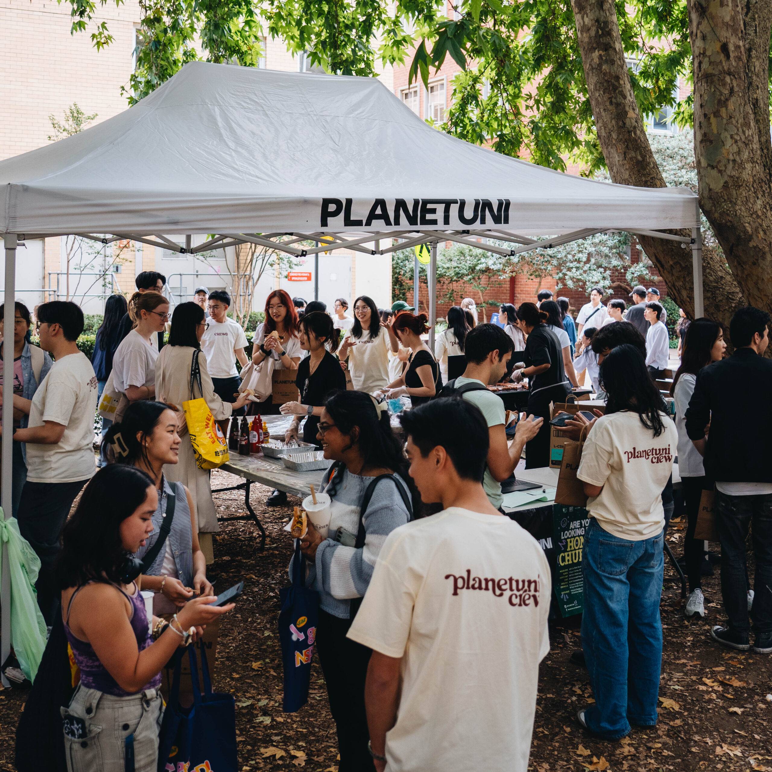 Group of people gathered under a white canopy at an outdoor campus fair, many wearing PlanetUni Crew shirts and handing out items.
