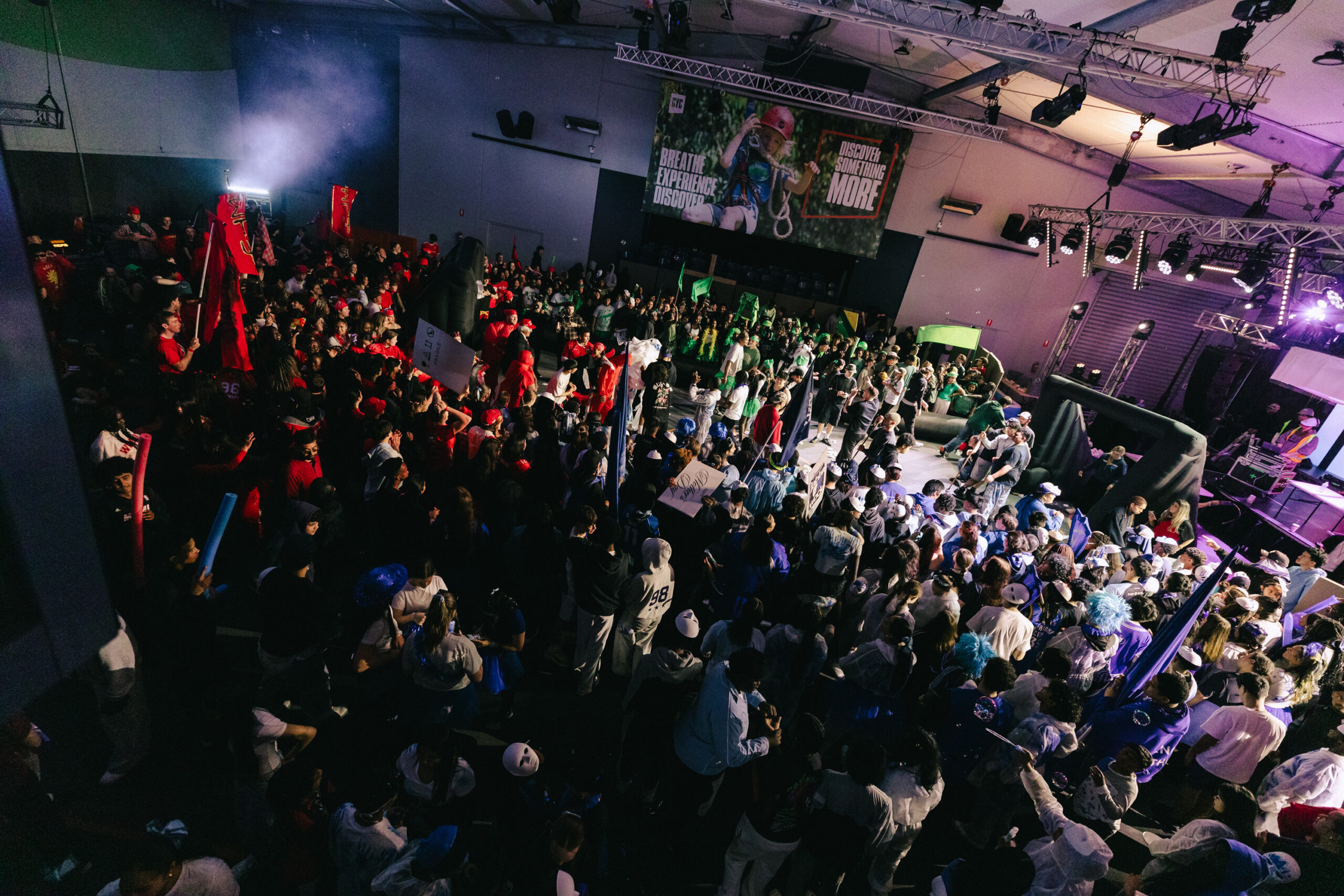 Large crowd gathered in an indoor arena for a live event, with stage lighting and rigging visible on the right.