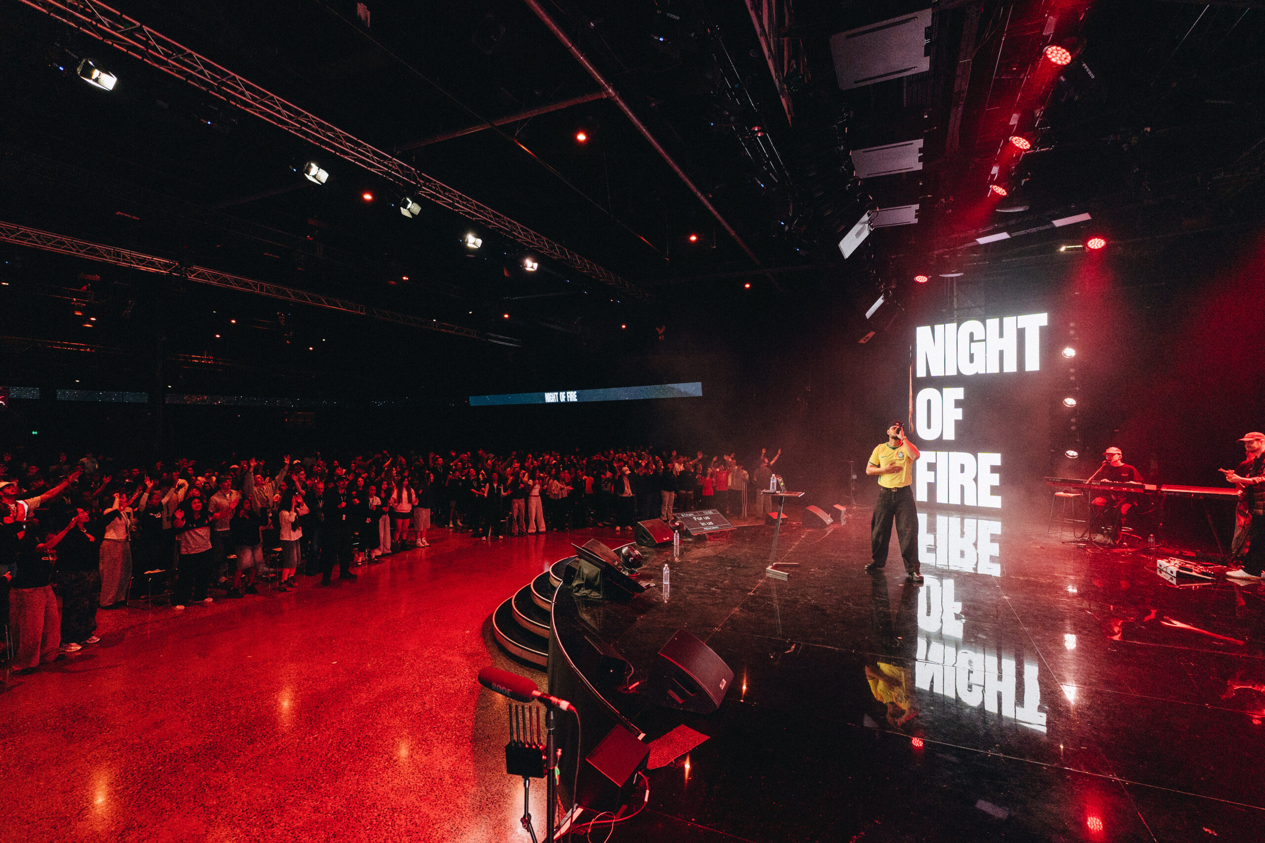 Performer on a dark stage under red lights with a tall vertical screen reading 'NIGHT OF FIRE' and a live band to the right, crowd visible in front.