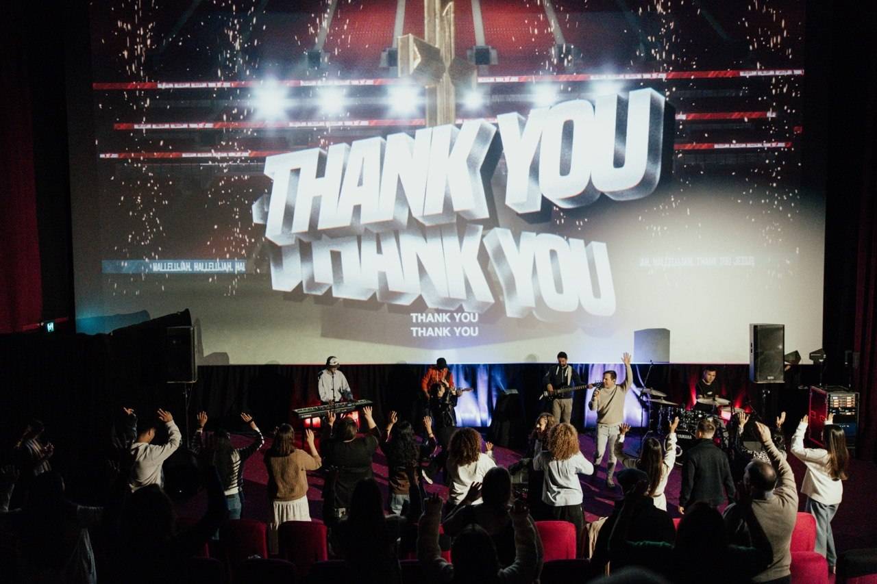 Band performs on a lit stage in a theater; a large screen behind reads 'THANK YOU' as the audience raises their hands in the foreground.